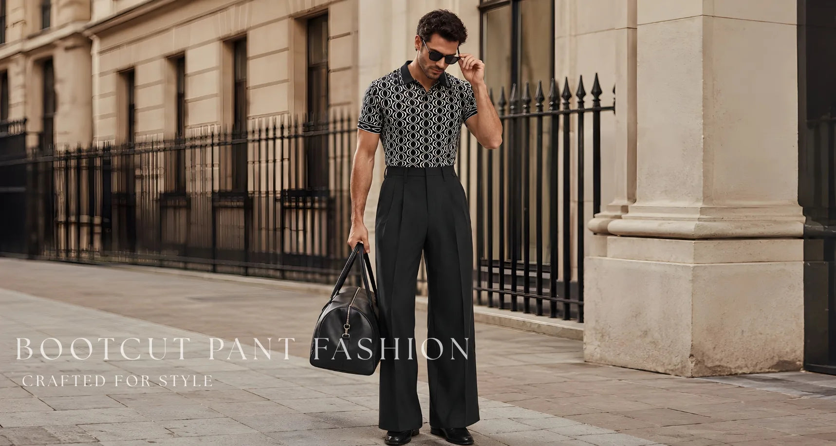 A man stands on a city sidewalk wearing a patterned short-sleeve button-up shirt and wide-leg dark trousers, holding a black tote bag. The background shows a beige stone building with a black metal fence.