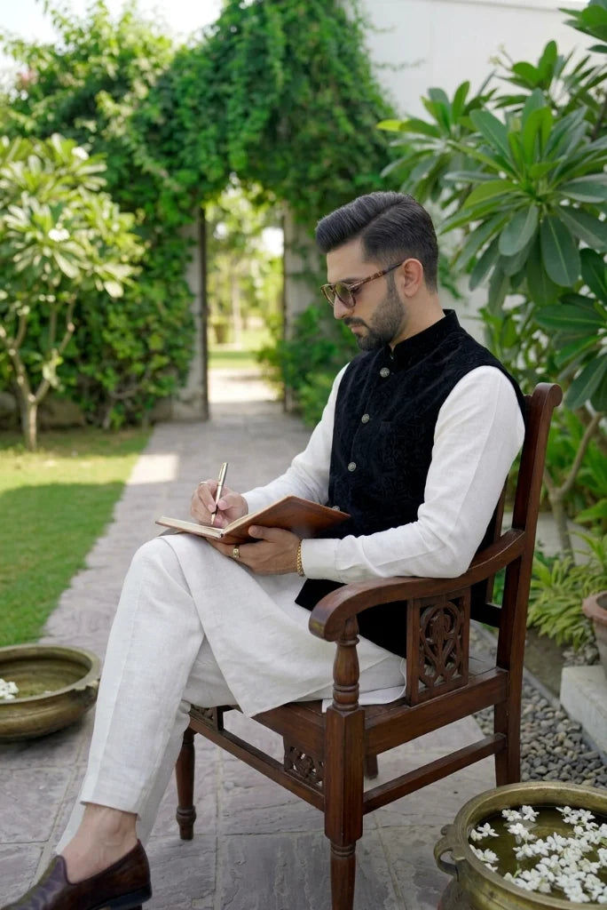 Man wearing black velvet Nehru collar waistcoat with golden buttons over white kurta and white trousers